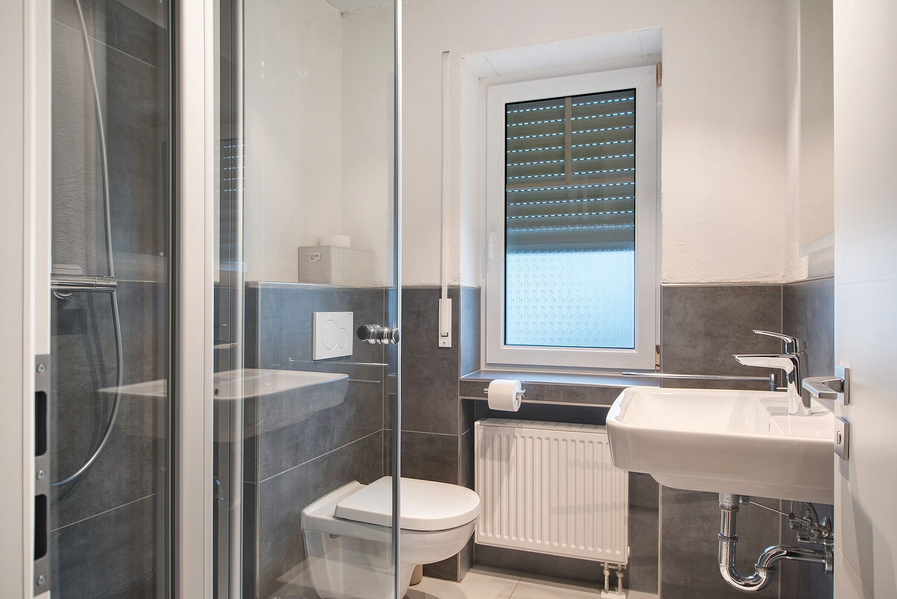 Un baño moderno con cabina de ducha de vidrio, inodoro, lavamanos y ventana con persianas.