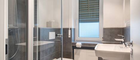 Un baño moderno con cabina de ducha de vidrio, inodoro, lavamanos y ventana con persianas.