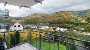 A balcony with a metal railing overlooking a residential area with houses and a hilly landscape.
