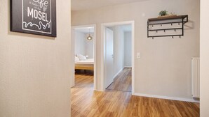 A hallway with a wooden floor, a bed in the background, a wall-mounted shelf with a plant, and a framed quote on the wall.