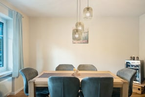 A dining area with a wooden table, four chairs, hanging pendant lights, and a refrigerator.