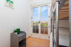 A compact room with a bunk bed, a small desk with a plant, and a sliding glass door leading to an outdoor balcony.