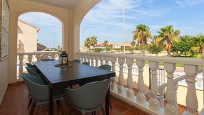 A covered outdoor dining area with a table and chairs, a railing, and a view of palm trees and buildings.