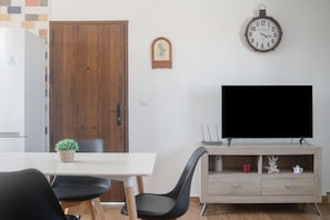 A modern living room with a white table, black chairs, a wooden door, a clock, and a TV on a wooden stand.