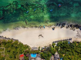 Vista aérea de una playa con aguas cristalinas, embarcaciones y un área de piscina con sombrillas.