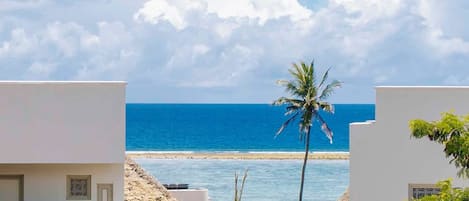 A coastal hotel with a view of the sea, palm trees, and a thatched-roof structure.