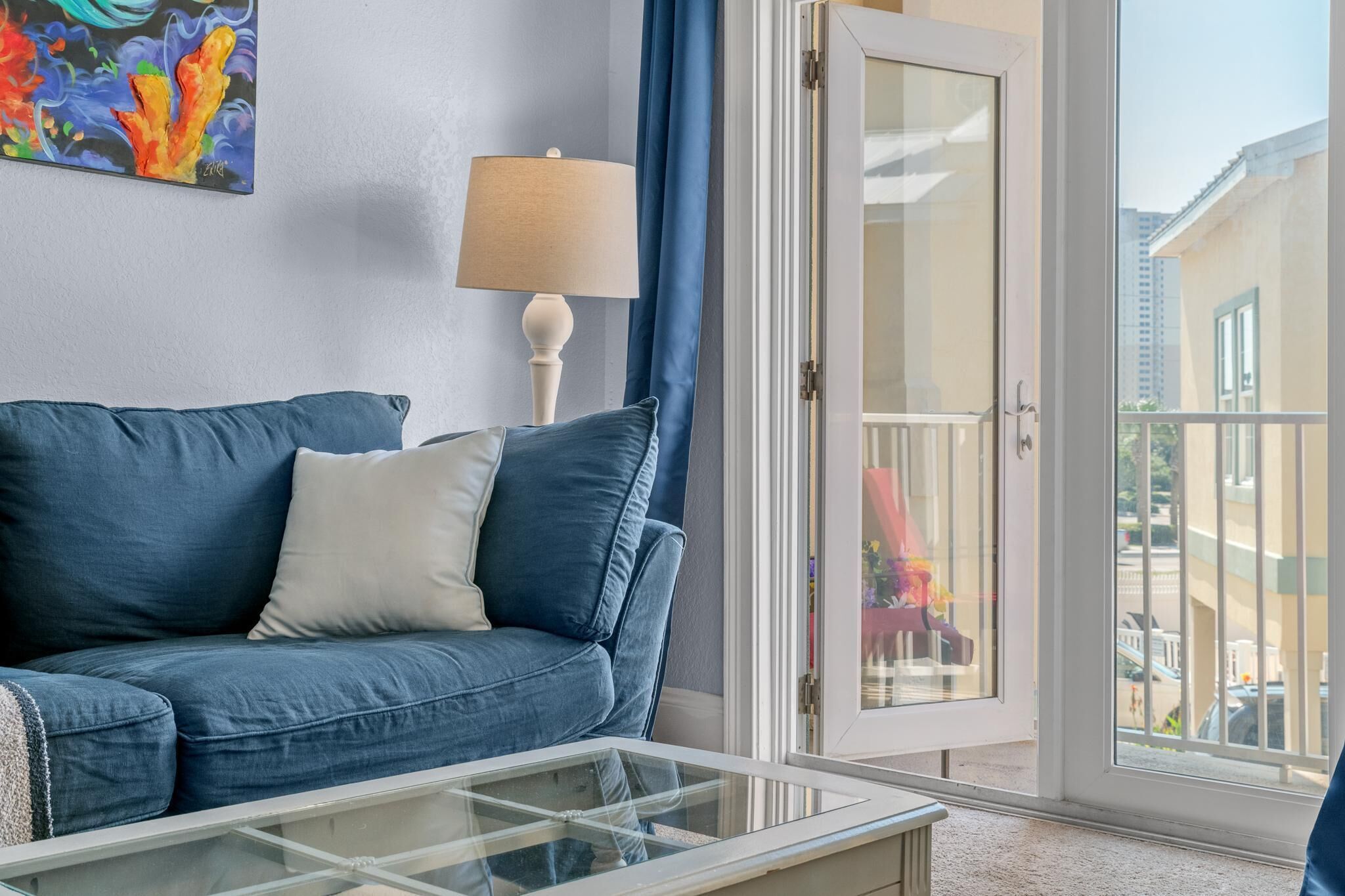 Una sala de estar con un sofá azul, una mesa de centro de vidrio, una lámpara y una puerta que da a un espacio al aire libre.