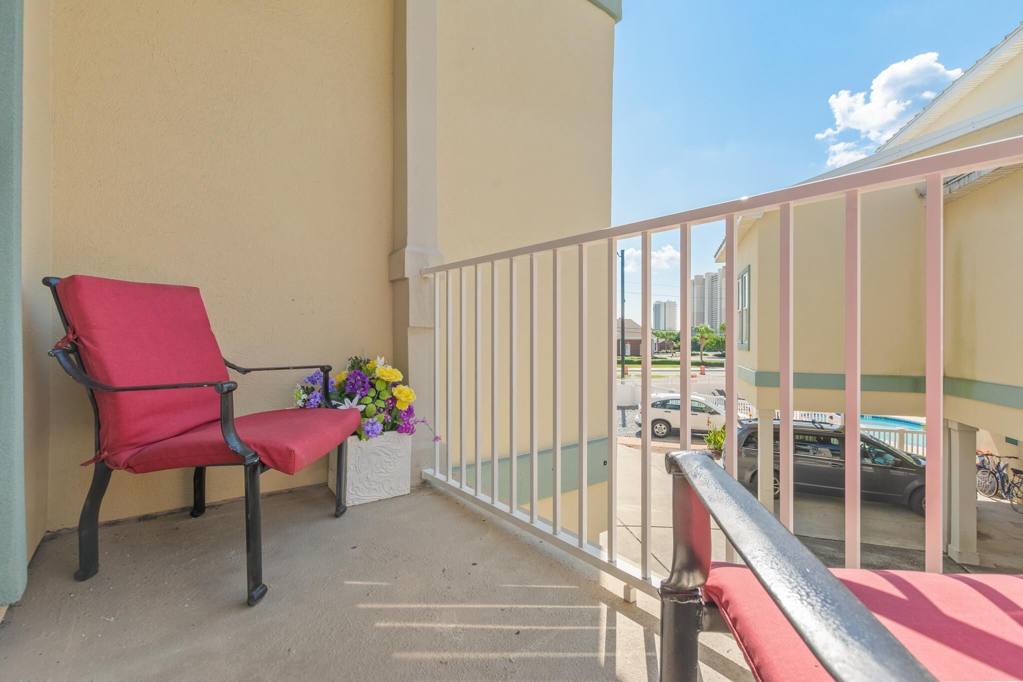 Un balcon avec une chaise rouge, un pot de fleurs blanc et une vue sur les voitures garées et les immeubles.