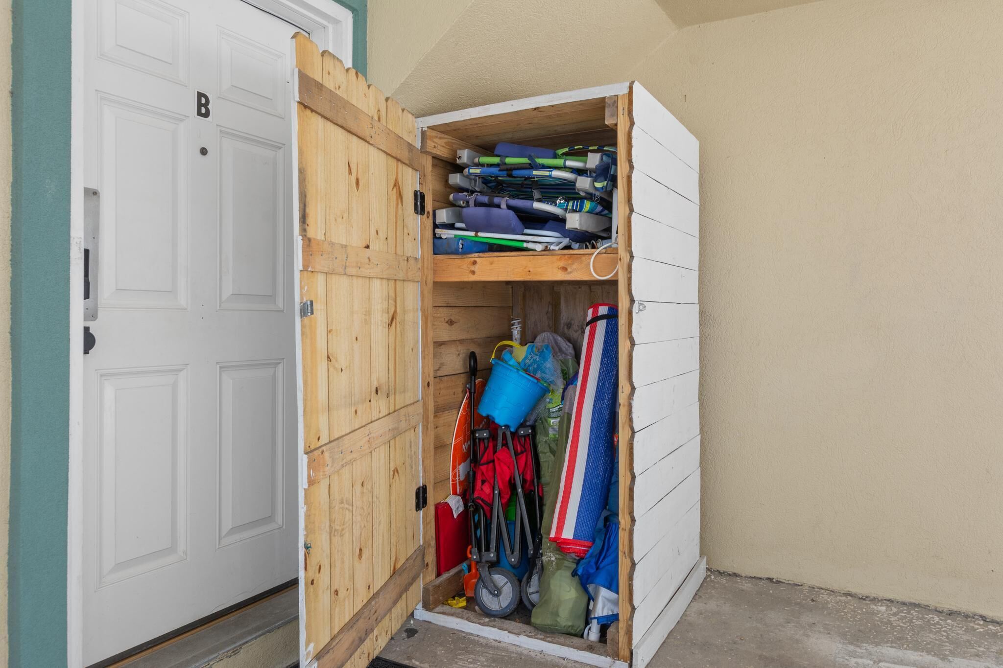 A storage unit with a wooden door, containing various items like a blue bucket, red and black paddles, and a striped towel.