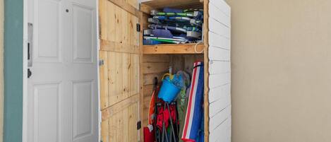 A storage unit with a wooden door, containing various items like a blue bucket, red and black paddles, and a striped towel.