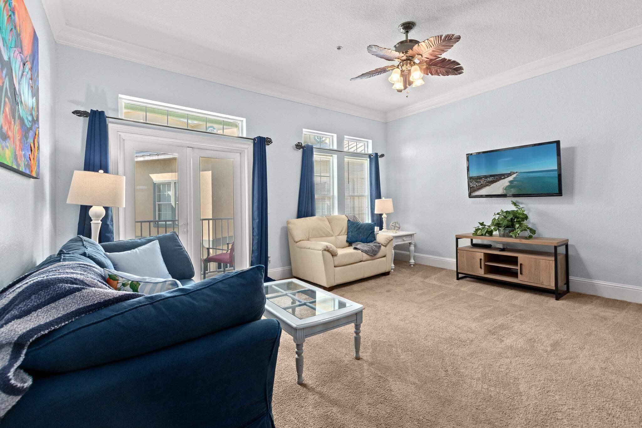 A living room with a blue sofa, beige armchair, and a coffee table. There is a TV mounted on the wall and a ceiling fan.
