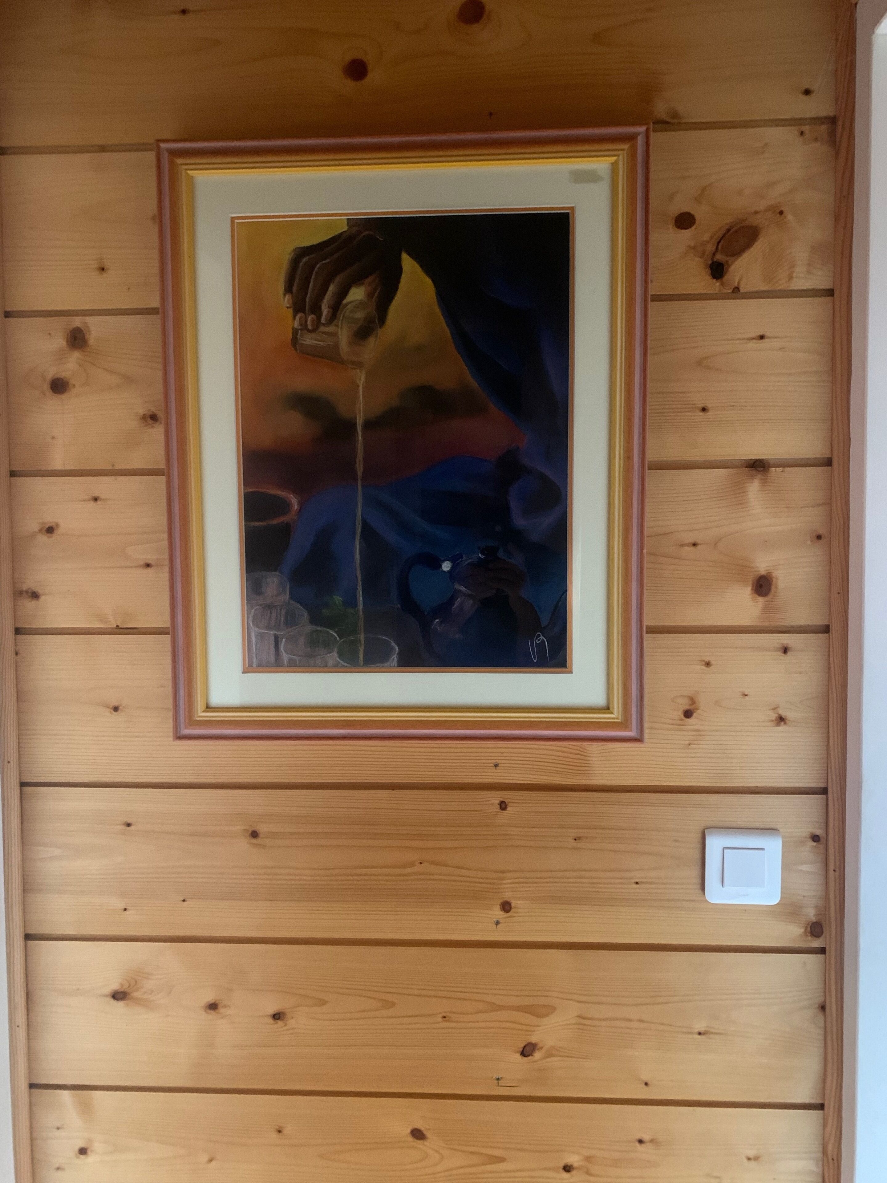 A framed painting of a hand pouring liquid, mounted on a wooden wall with a light switch below it.