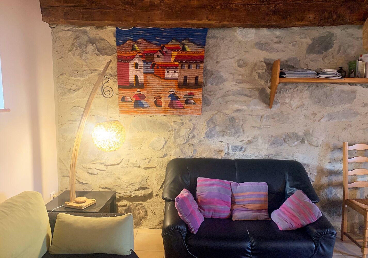Una sala de estar acogedora con pared de piedra, un sofá de cuero negro con cojines coloridos, una mesita con una lámpara y un cuadro de un paisaje campestre.