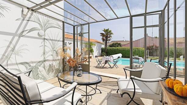 Un invernadero con paredes de vidrio, una piscina, muebles blancos y un mural tropical.