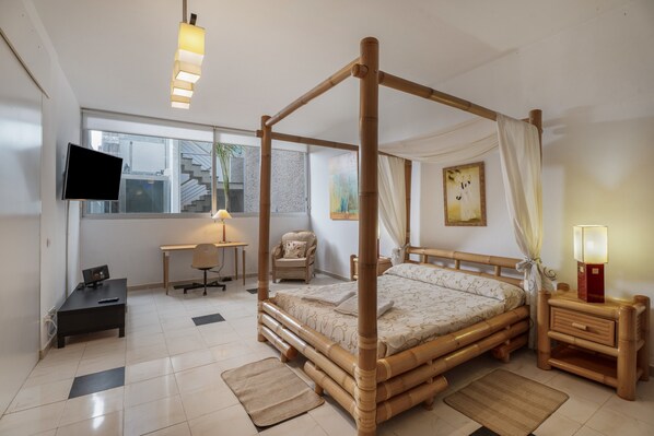 A room with a four-poster bed, a desk with a chair, a television, and a bedside table with a lamp.