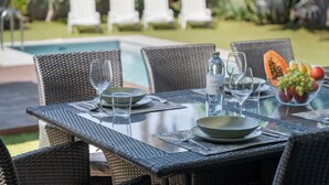 An outdoor dining area with wicker chairs and a glass table, a pool in the background, and a fruit bowl on the table. - Vacation Villa 'Playa Del Duque' with Private Terrace, Wi-Fi and Air Conditioning (Adeje)