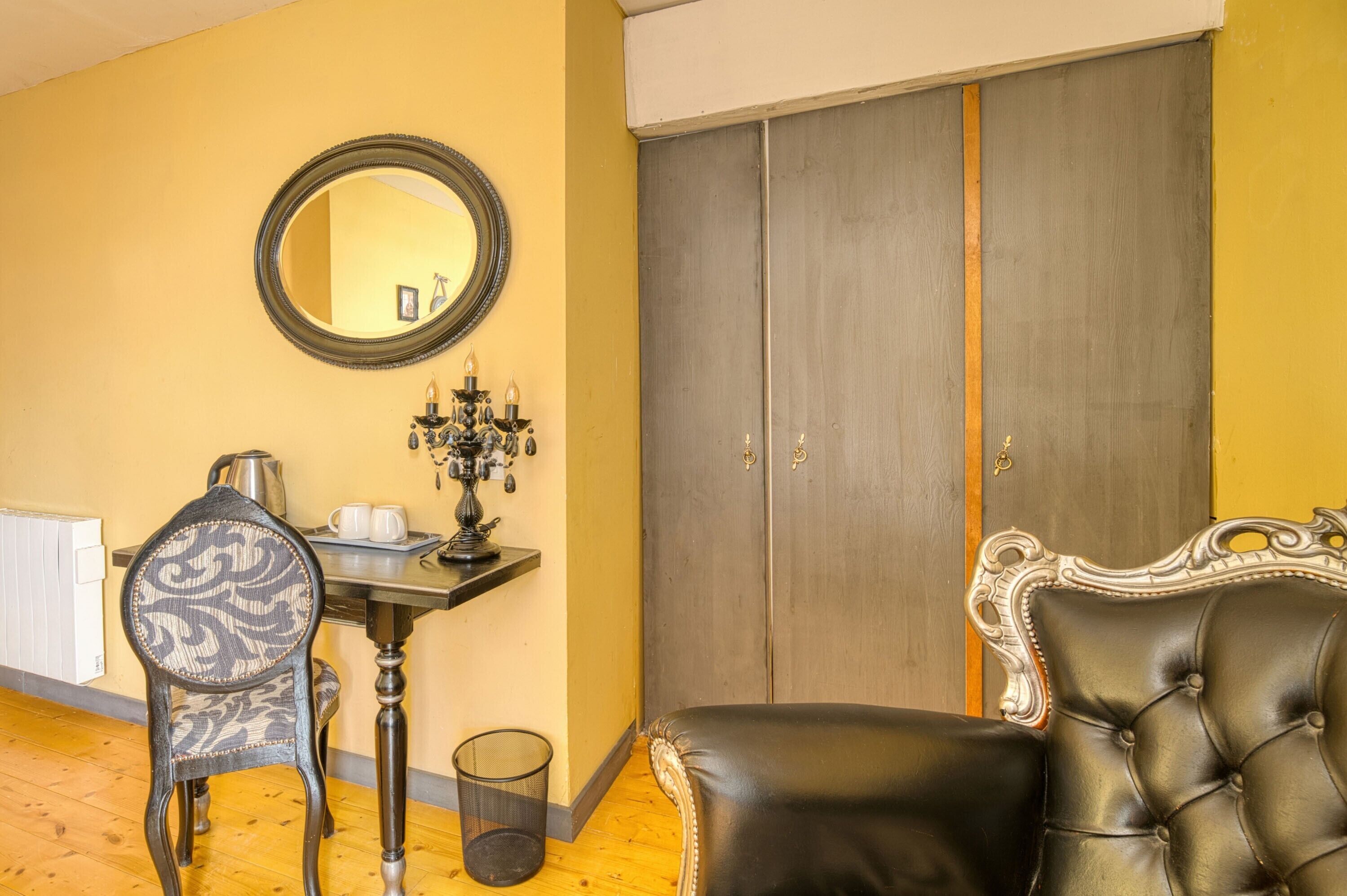 A room with a wooden floor, a round mirror, a dark wooden table with a candelabra, a black leather chair, and a large wardrobe.