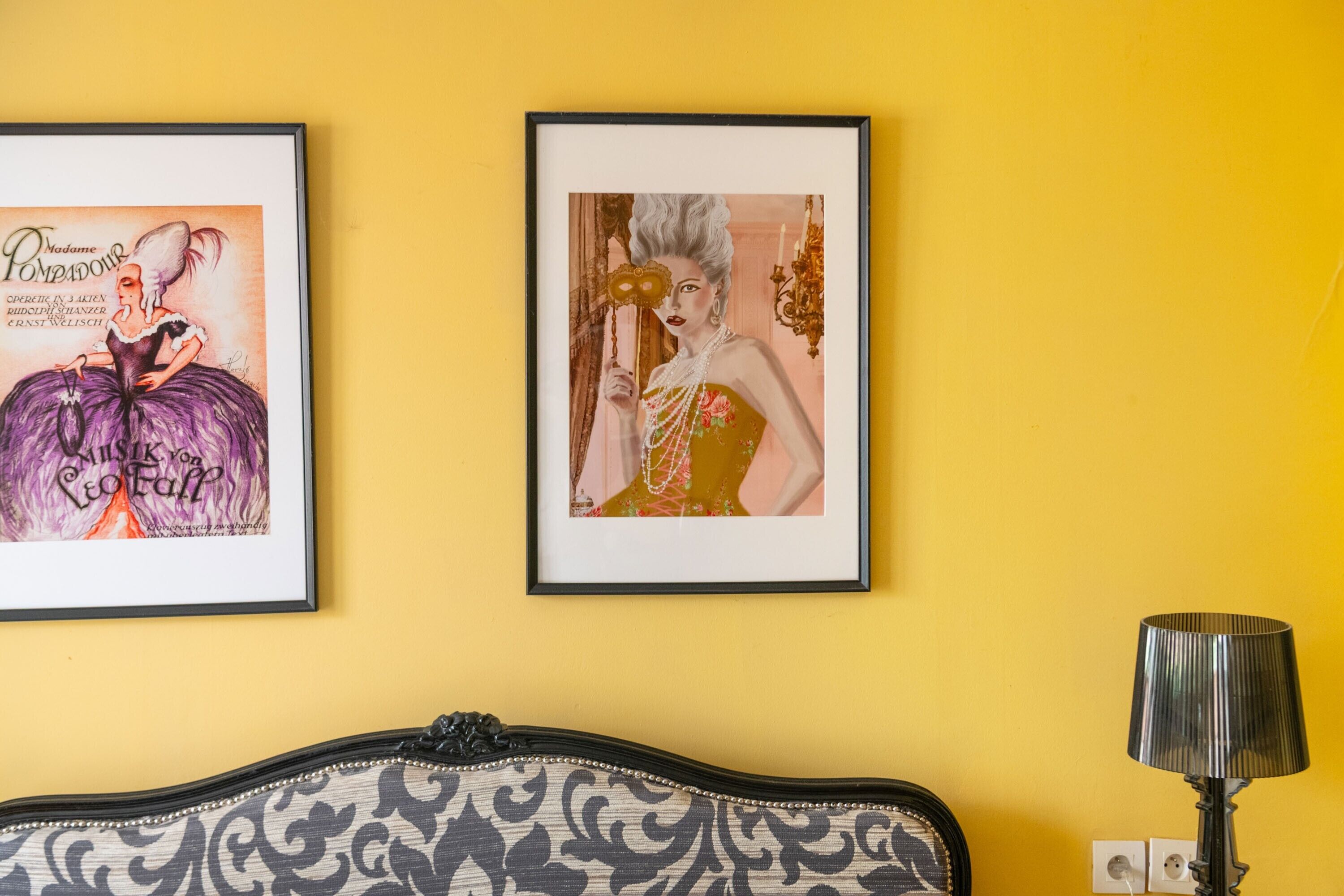 Twee ingelijste kunstwerken aan een gele muur, een zwarte lamp en een gestreept sofa.