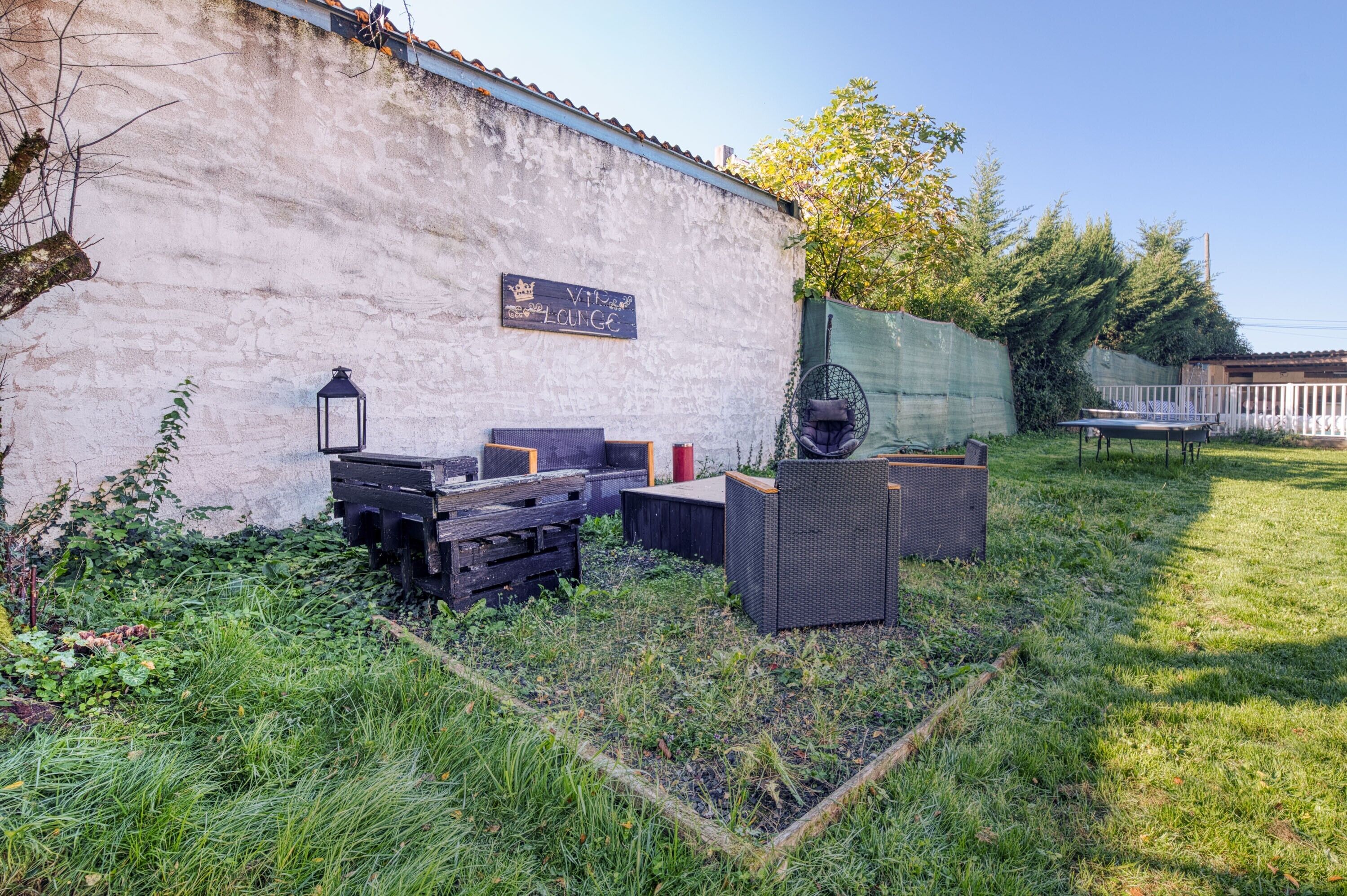 Un espace extérieur aménagé dans un style rustique, avec une pancarte indiquant « Vine Lounge », entouré de verdure et délimité par une clôture blanche.