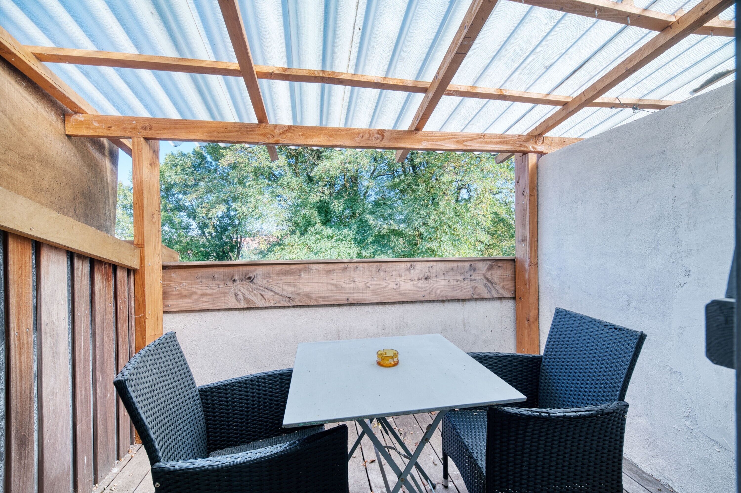 A covered patio with a table and two chairs, wooden beams, and a view of trees.