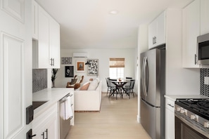 Una cocina moderna con armarios blancos, un refrigerador de acero inoxidable y una estufa a gas. Hay un comedor con una mesa y sillas, y un sofá al fondo.