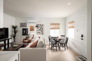 Una sala de estar moderna con un sofá, una mesa de centro y un televisor. Cuenta con un comedor con mesa y sillas. La habitación tiene obras de arte en las paredes y una ventana con persianas.