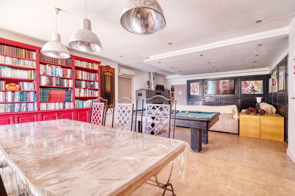 A spacious room with a pool table, a dining table, and a large bookshelf filled with books.
