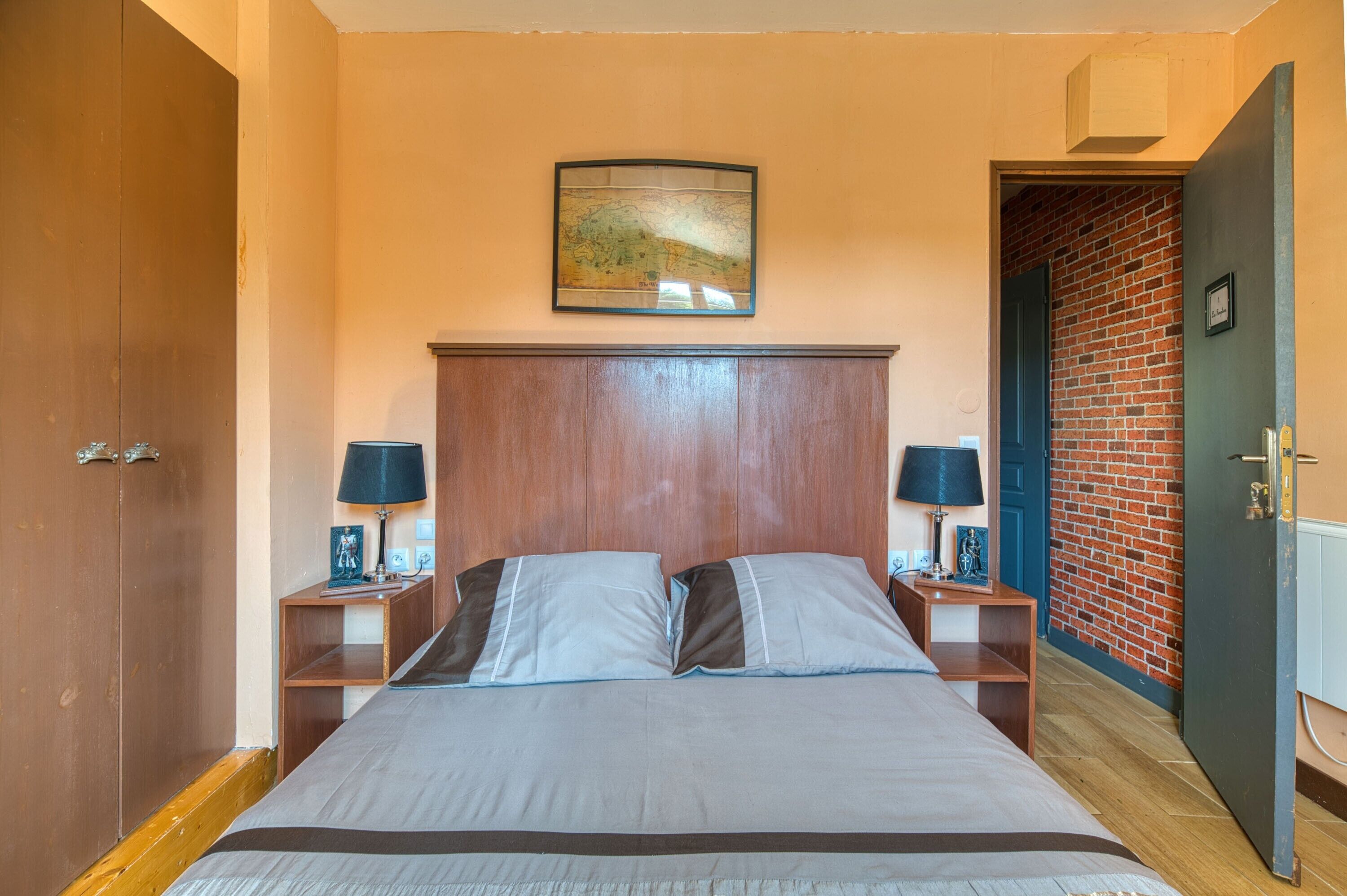 A bedroom with a wooden headboard, two bedside tables with lamps, a bed with grey bedding, a map on the wall, and a brick wall in the background.