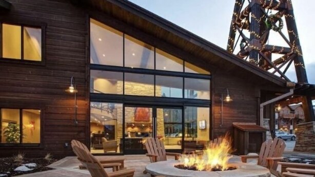 A modern building with large windows, a stone fire pit, and wooden chairs.
