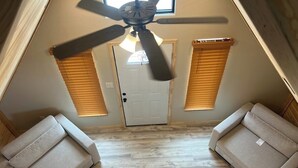 A room with a sloped ceiling, a ceiling fan, two sofas, a door, and a window with blinds.