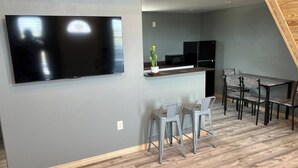 A modern kitchen with a flat-screen TV, grey walls, and a dining area with metal chairs.
