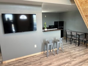A modern kitchen with a flat-screen TV, grey walls, and a dining area with metal chairs.