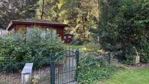 A rustic cabin surrounded by dense foliage, with a metal gate and a solar panel in the foreground. - Chaletpark Kempenbos - 196 (Diessen)