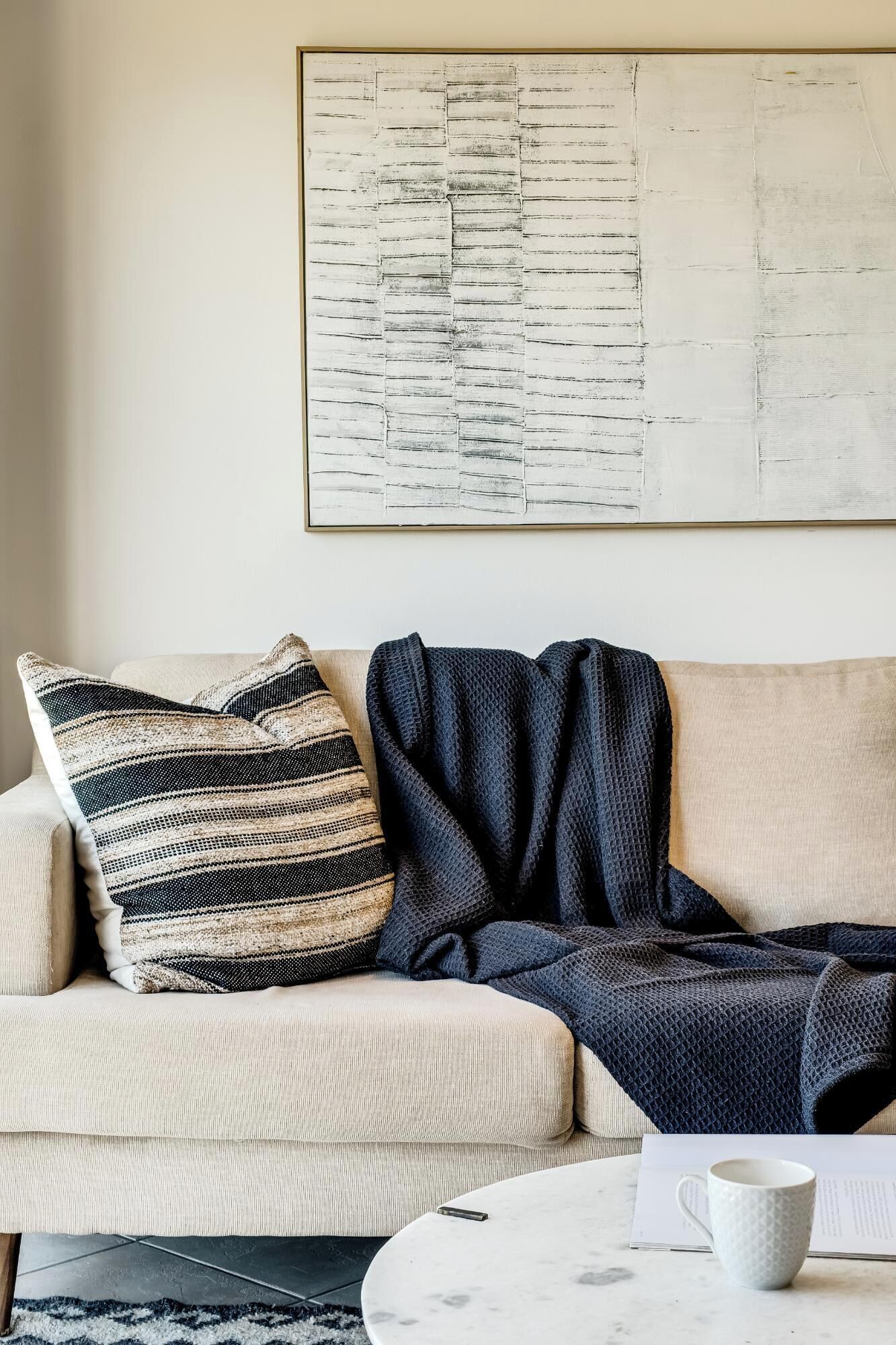 En stue med en beige sofa, en pude med et mønster, et mørkeblåt tæppe og et hvidt sofabord med en kop.