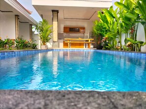 A swimming pool with a wooden deck, surrounded by tropical plants. - Alit Guest House (Jimbaran)