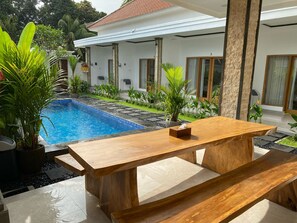 A poolside area with a wooden bench, a small table, and a house in the background. - Alit Guest House (Jimbaran)