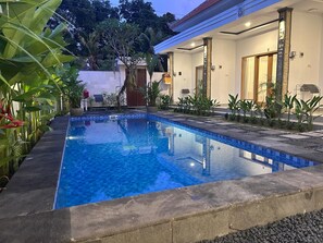 A swimming pool with a tiled edge, surrounded by plants and a building with a tiled roof. - Alit Guest House (Jimbaran)