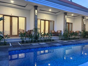 A poolside area with a tiled pool, wooden chairs, and potted plants. - Alit Guest House (Jimbaran)