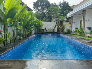 A rectangular swimming pool with a tiled edge, surrounded by lush green plants and a white building with columns. - Alit Guest House (Jimbaran)