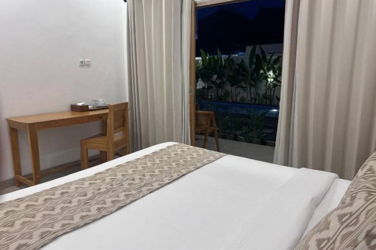 Ein Hotelzimmer mit Bett, Schreibtisch und Stuhl. Durch das Fenster ist eine Aussicht auf Pflanzen und ein Gebäude zu sehen.