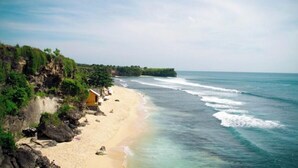Uma praia com um pequeno prédio amarelo, água azul cristalina e um penhasco rochoso coberto de vegetação.
