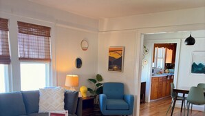 Ein Wohnzimmer mit Sofa, Couchtisch und einem Sessel. Es gibt einen Essbereich mit Tisch und Stühlen. Im Hintergrund ist eine Küche erkennbar.