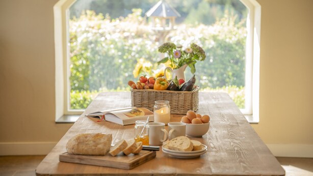 Een ontbijt opstelling met een rieten mand vol fruit, eieren, brood, en een kaars op een houten tafel bij een raam met uitzicht op groen.