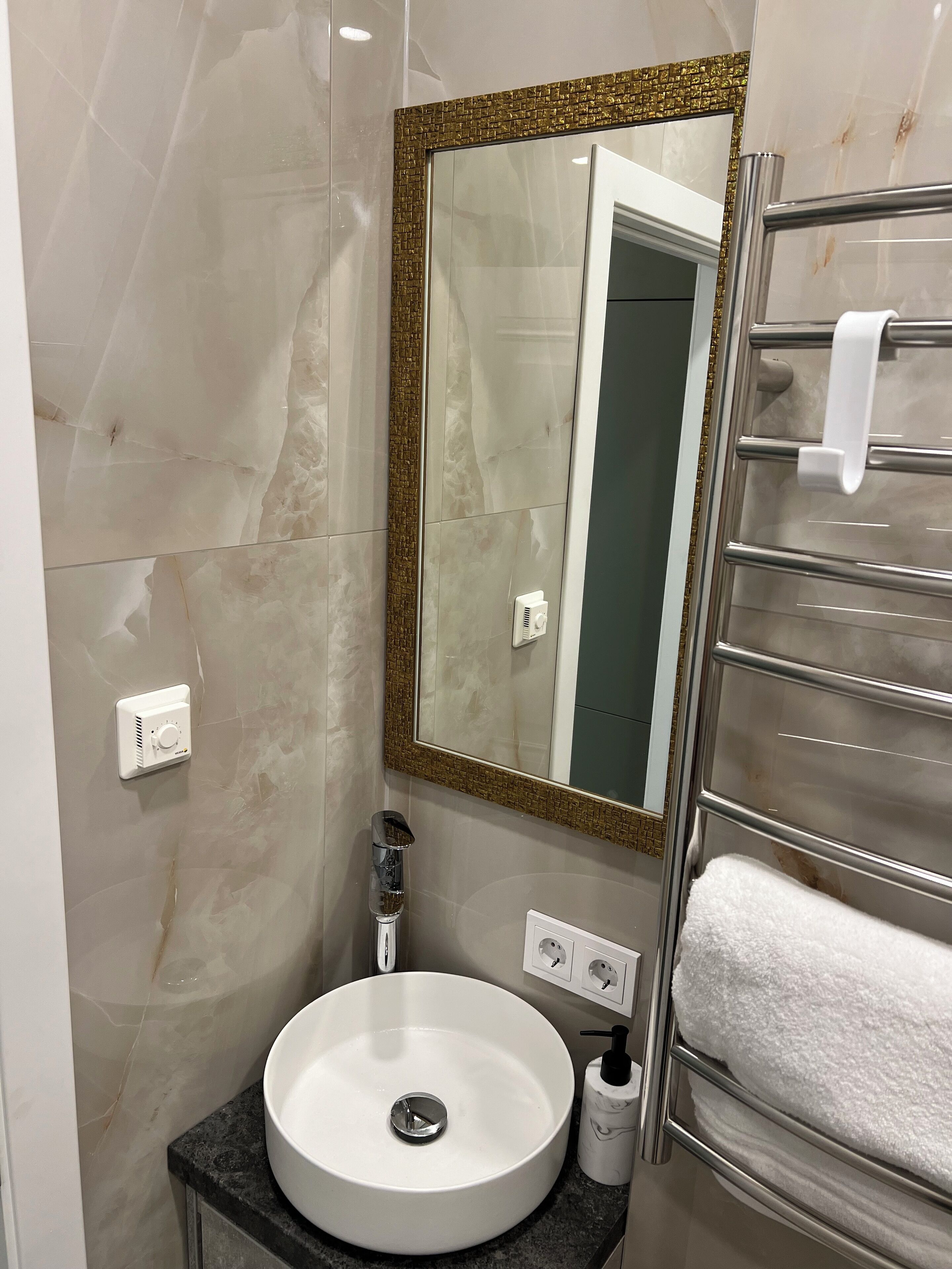 A bathroom with a white sink, a mirror with a gold frame, a towel rack, and a bottle of hand soap.