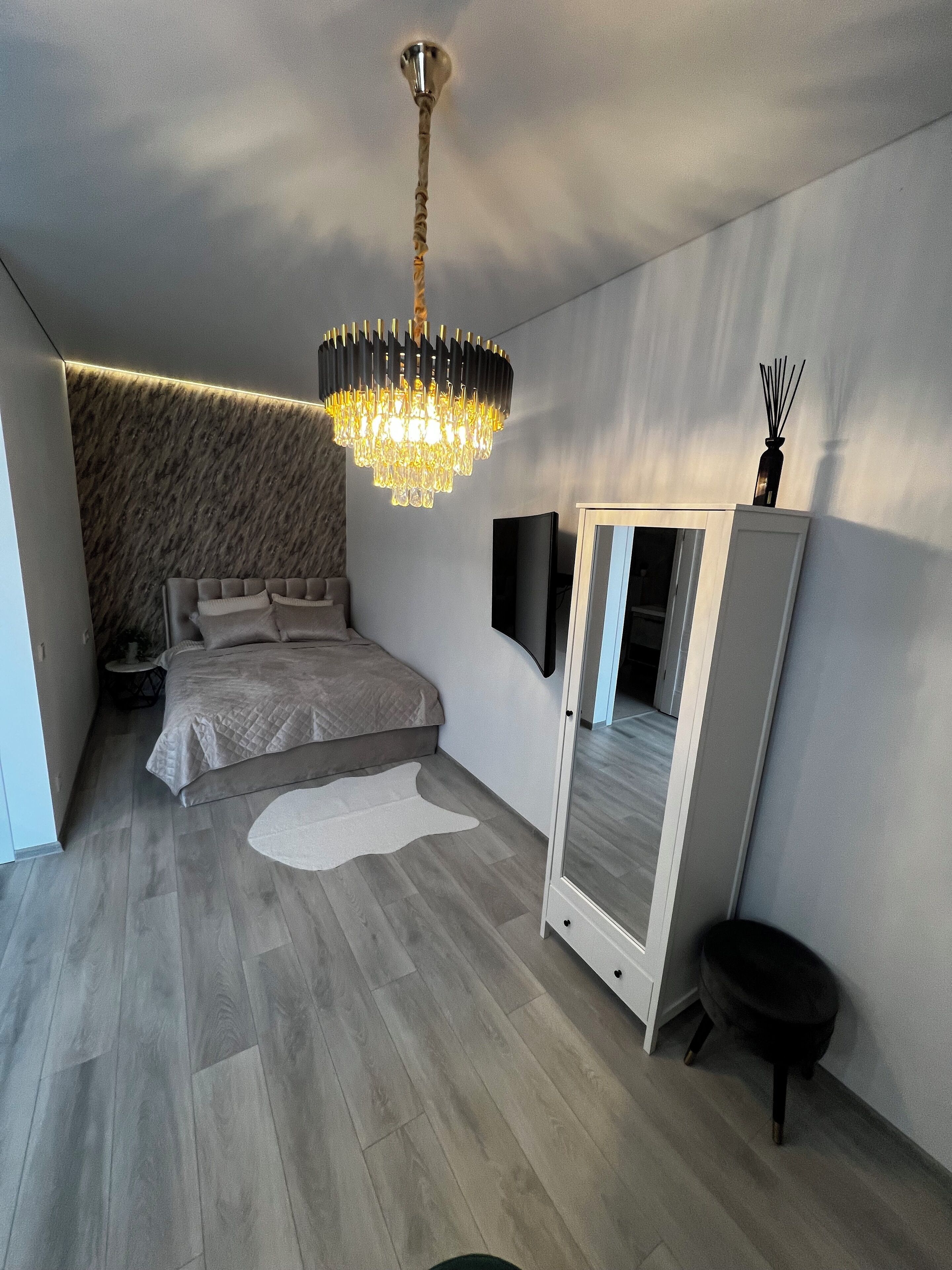 A modern bedroom with a large bed, a chandelier, a wardrobe, and a mirror.