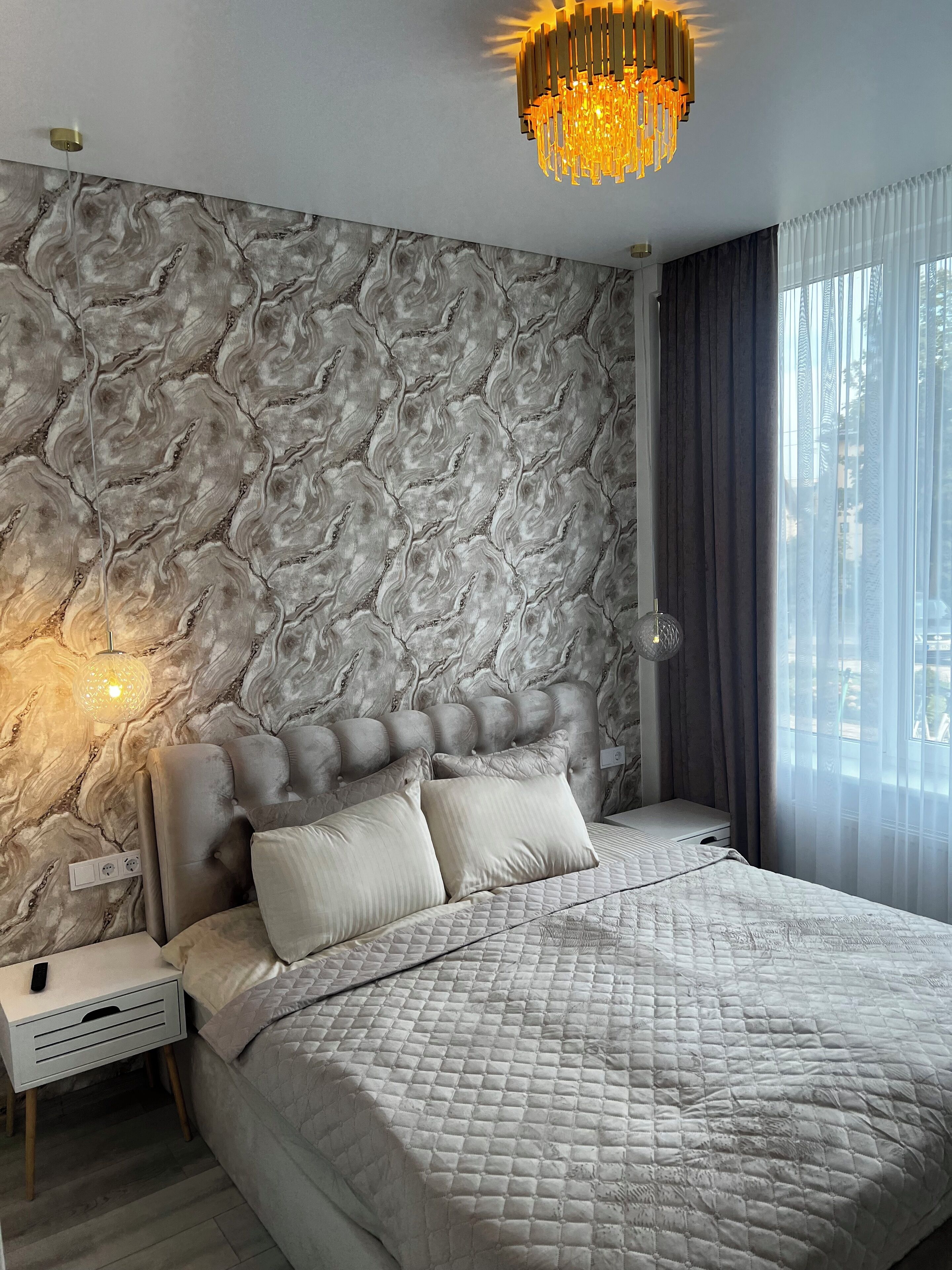 A bedroom with a marble-patterned wall, a tufted headboard, a quilted bedspread, and a chandelier.