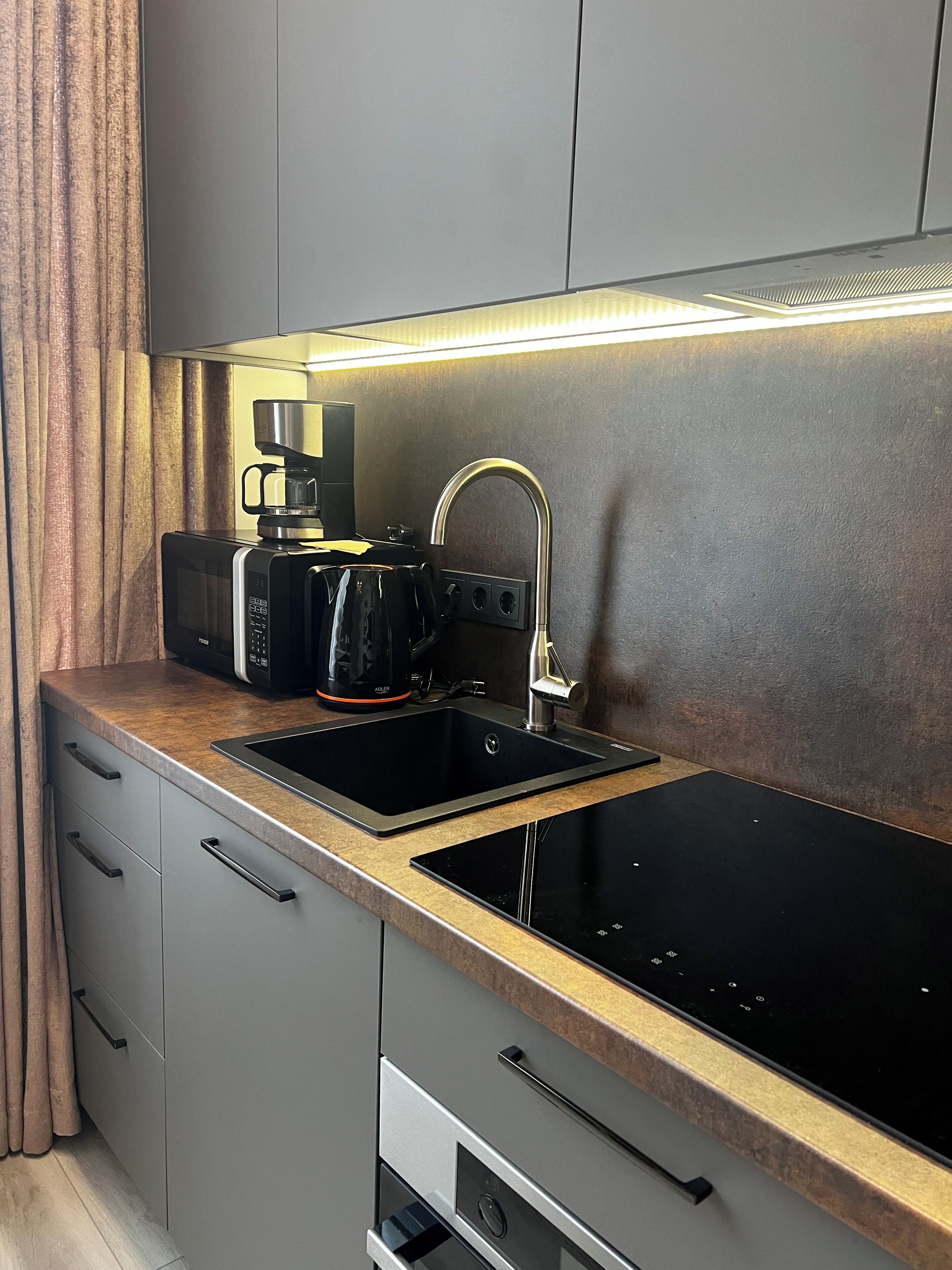 A modern kitchen with a black induction cooktop, a built-in oven, a coffee maker, and a stainless steel sink.