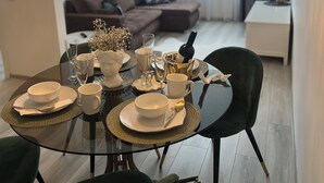 Ein Essbereich mit einem runden Glastisch, gedeckt für vier Personen, umgeben von grünen, gepolsterten Stühlen. Eine Pendelleuchte hängt darüber, und ein Sofa ist im Hintergrund zu sehen.