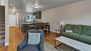 A modern living room with a green sofa, a blue armchair, and a coffee table. There is a dining area with a wooden table and chairs. The kitchen is visible with stainless steel appliances. - Flexible Extended Stay 3BR - Spacious + Fireplace + Patio (Salem)