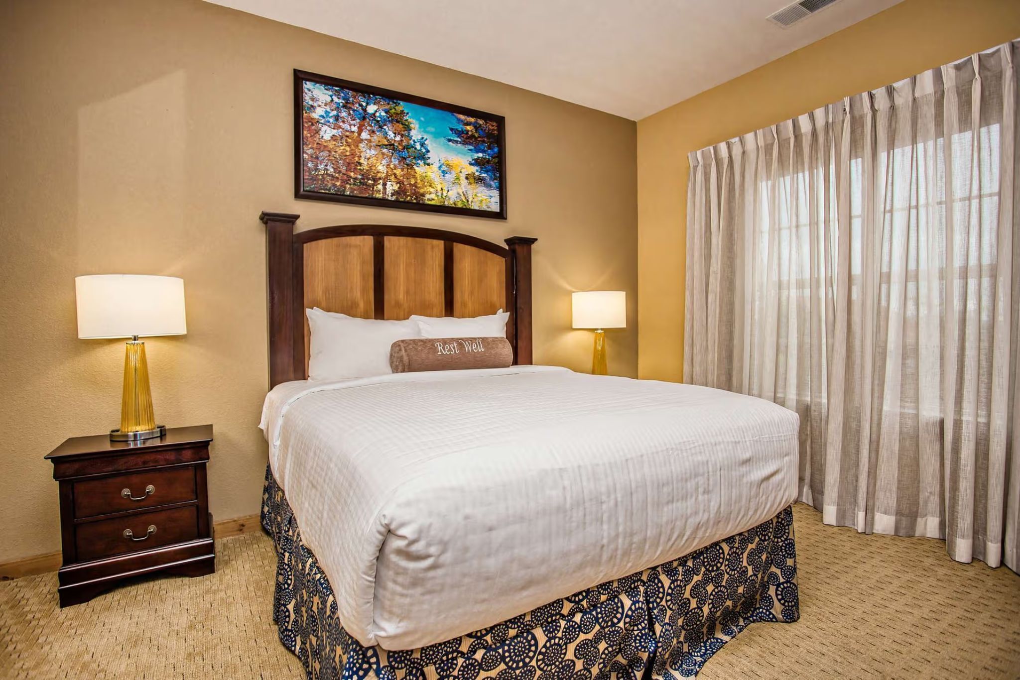 Habitación de hotel con una cama grande, dos lámparas de noche, un cuadro en la pared y una ventana con cortinas.